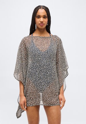 Femme aux cheveux tressés portant une tunique transparente à imprimé léopard par-dessus un maillot de bain noir, debout devant un fond clair uni.