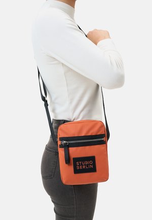 Orange farbene Nylon-Crossbody-Tasche mit einem schwarzen Reißverschluss und einem schwarzen Etikett, auf dem "STUDIO BERLIN" steht. Verfügt über einen verstellbaren Riemen und eine rechteckige Form.