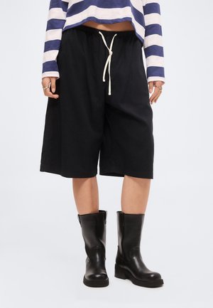 Person iført sorte knælange shorts med snor, sorte læderstøvler til midt på læggen og en blå- og hvidstribet langærmet top.