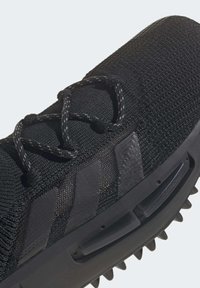 Svart atletisk sko gjord av texturerat stickat material. Utrustad med svarta snörningar, tre randiga accenter och en spikad svart sula.