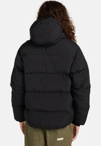 Sort pufferjakke med hætte, der har lodret quiltede sektioner, et struktureret stof og en lynlåslukning. Bagsiden viser et minimalistisk design.