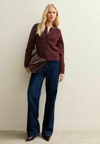 Burgundowy sweter kardigan z ozdobnym kołnierzem, zestawiony z ciemnoniebieskimi dżinsami w stylu dzwonów oraz błyszczącą bordową kopertówką. Szpiczaste bordowe szpilki dopełniają całość.