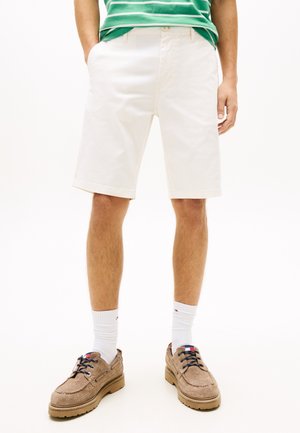 Hombre vestido con pantalones cortos blancos hasta la rodilla, camisa verde con rayas, calcetines blancos hasta el tobillo y zapatos casuales marrón claro con cordones, de pie con las manos en los bolsillos.