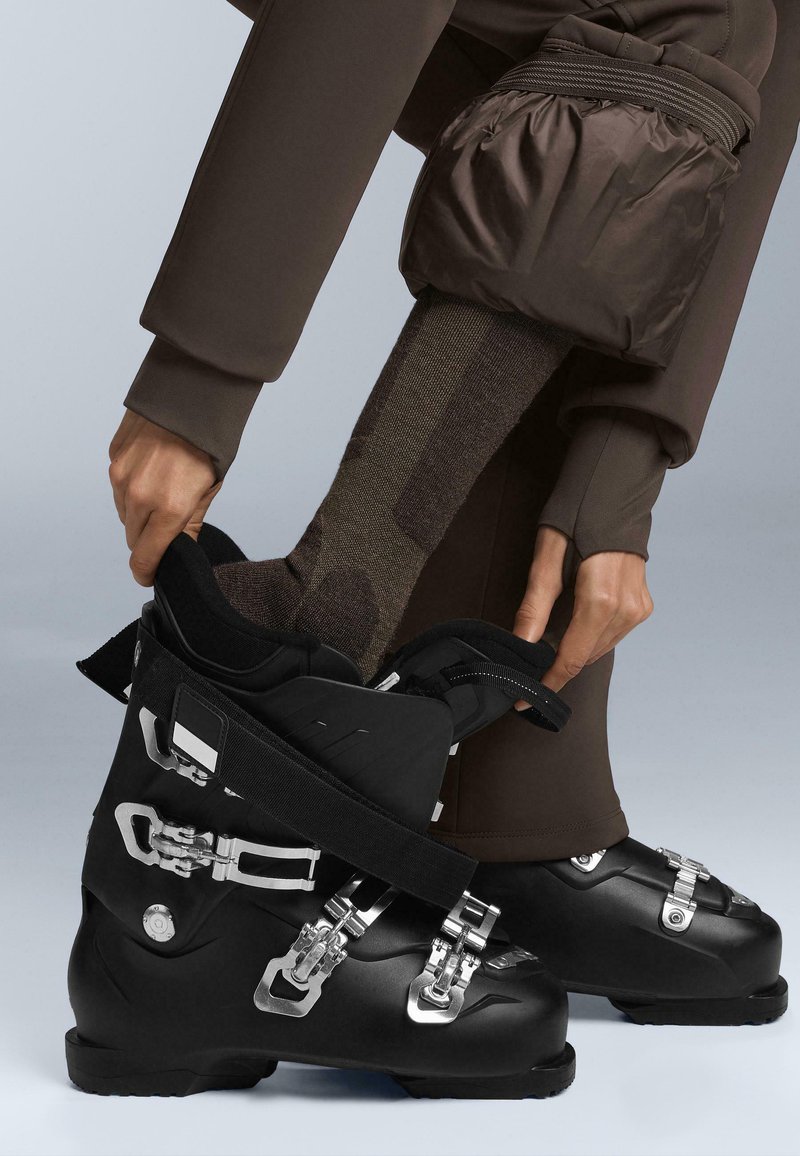 Black ski boots with silver metallic buckles and straps, featuring a durable plastic design, are being fitted with a patterned sock visible above.