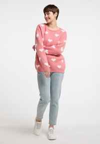 Rosa Pullover mit weißen Herzmustern, Rundhalsausschnitt und langen Ärmeln, kombiniert mit hellblauen Jeans und weißen Sneakern.