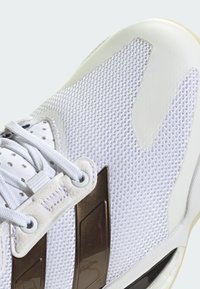 Weiße Mesh-Sneaker mit strukturiertem Strick-Obermaterial, metallisch braunen Akzenten, Kunststoff-Ösen und einer Gummisohle in einem schlanken Design.