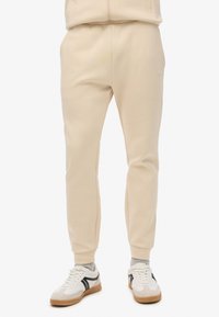 Sudadera beige con un ajuste tapered, hecha de un tejido suave. Presenta una cinturilla elástica y puños acanalados. Combinada con zapatillas blancas.