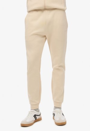 Sudadera beige con un ajuste tapered, hecha de un tejido suave. Presenta una cinturilla elástica y puños acanalados. Combinada con zapatillas blancas.