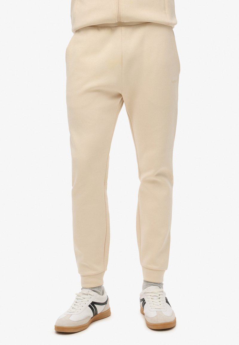 Sudadera beige con un ajuste tapered, hecha de un tejido suave. Presenta una cinturilla elástica y puños acanalados. Combinada con zapatillas blancas.