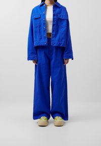 Blå denimjacka med två bröstfickor och gröna detaljer, tillsammans med vida blå denimbyxor och gröna sneakers med beigefärgade sulor.