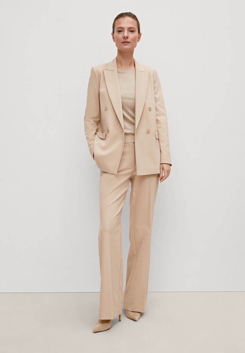 Blazer beige à double boutonnage avec revers en notch, associé à un pantalon assorti et un haut beige. Tissu lisse, silhouette ajustée, accents minimalistes.