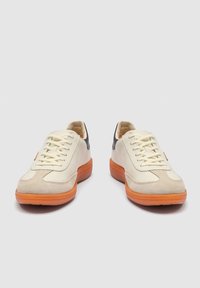 Pari kermaan värisiä nahkakenkiä, joissa on beigeä mokkanahkaa varvassuojissa, mustat kantakappaleet, valkoiset nauhat ja oranssit kumipohjat harmaalla taustalla.