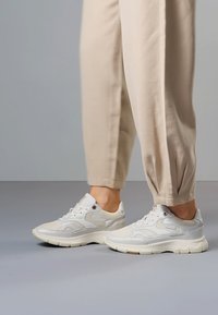 Piedi con indosso sneakers bianche chunky abbinate a pantaloni beige a gamba stretta su uno sfondo grigio neutro.