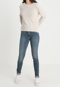 Maglione a maglia beige chiaro con scollo a girocollo, abbinato a jeans blu slim-fit e sneakers bianche. Tessuto testurizzato con dettagli a coste.