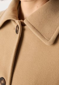 Gros plan sur le col d'un manteau en laine beige avec des boutons marron foncé, porté par une personne dont le cou et le menton sont visibles.