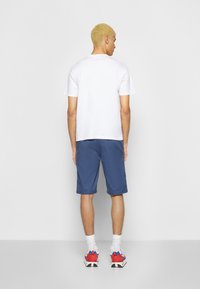 T-shirt de algodão branco, calções azuis e sapatilhas atléticas vermelhas com detalhes em azul; visto de trás, destacando os pormenores das roupas e calçado.