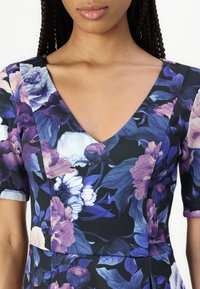 Virágmintás ruha mély V-nyakkivágással, sötét anyagból, lila és fehér virágokkal, rövid ujjakkal és szűkített felsőrésszel.