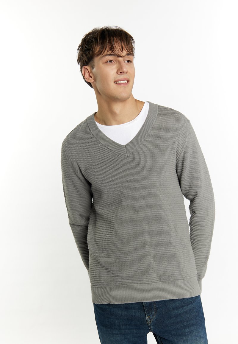 Maglione grigio a V con texture, con polsini e orlo a costina, indossato sopra una t-shirt bianca, abbinato a jeans blu.