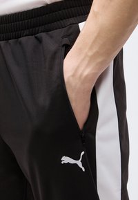 Χέρι στην τσέπη μαύρου φόρμας Puma με λευκή πλευρική ρίγα και ορατό φερμουάρ στην τσέπη.