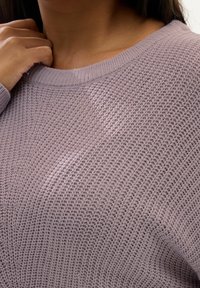 Helle lila Strickjacke aus strukturiertem Stoff, mit weitem Halsausschnitt und gerippten Bündchen. Nahaufnahme des Stoffmusters und der detaillierten Naht.