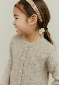 Vaaleanharmaa neulottu neuletakki, jossa on tekstuuri pinta, pyöreä kaulus ja pienet napit. Koristeltu kimaltelevalla hiuspannalla.