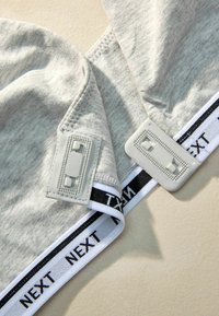 Šedá textilná podprsenka s nastaviteľnými ramienkami. Obsahuje biely pás s čiernym nápisom "NEXT" a šedými kovovými akcentmi na ramienkach.