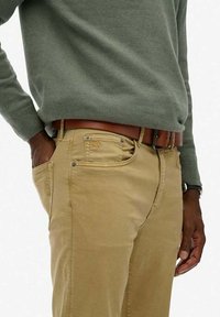 Chinos de algodón en color beige con corte recto, diseño de cinco bolsillos, remaches de metal y un cinturón de cuero marrón, combinados con un suéter de lana verde.