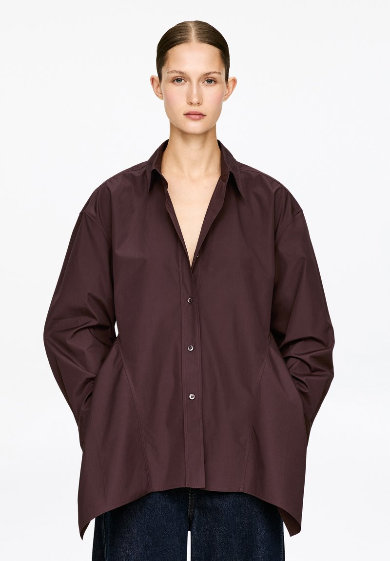 Chemise bordeaux à boutons avec une coupe ample, grand col, et détails sur les coutures latérales, fabriquée dans un tissu lisse. Associée à un denim foncé.