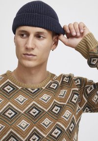 Bruine gebreide trui met diamanten in wit, tan en groen. Model draagt een marineblauwe geribbelde beanie, die ze met één hand vasthoudt.
