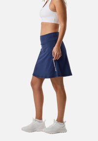 Mujer con sujetador deportivo blanco, falda deportiva azul marino y zapatillas de correr blancas, de pie de perfil sobre un fondo liso.