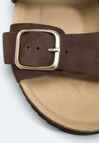 Bride de sandale en daim marron avec une boucle carrée dorée sur une semelle beige, présentée en gros plan.