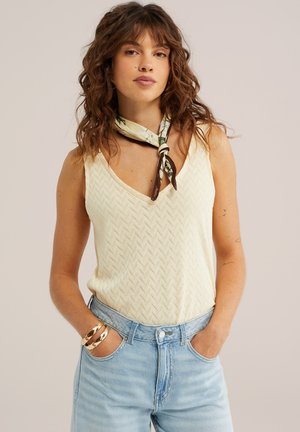 Mujer con cabello rizado que lleva un top de punto sin mangas color crema, vaqueros azules de talle alto, un pañuelo estampado en el cuello y pulseras doradas, con las manos en los bolsillos.