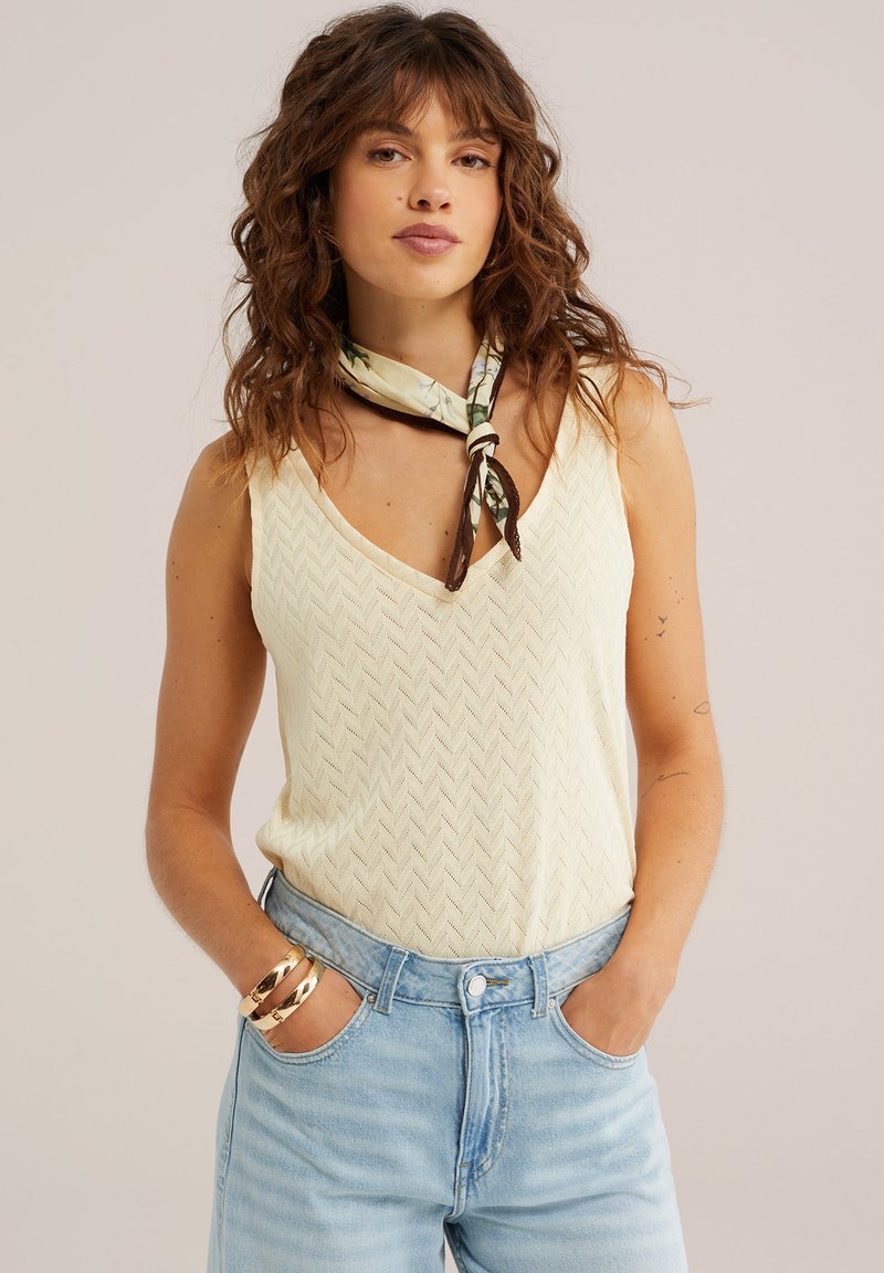 Femme aux cheveux bouclés portant un top en maille sans manches crème, un jean bleu taille haute, un foulard à motifs autour du cou et des bracelets dorés, les mains dans les poches.