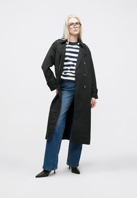 Cappotto trench nero con bottoni doppi, abbinato a una camicia a righe blu e bianca, jeans blu a gamba larga e stivali neri con tacco.