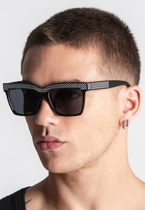 Occhiali da sole neri con una fascia decorativa a puntini neri e bianchi lungo la parte superiore. Lenti scure e un design di montatura robusto.