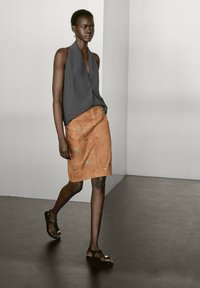 Top noir à dos nu, jupe en daim marron avec broderies florales et sandales noires. Modèle marchant sur un sol sombre devant un fond neutre.
