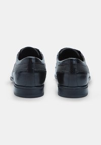 Un paio di scarpe da uomo eleganti in pelle nera viste da dietro, con dettagli testurizzati sopra il tacco e tacchi bassi impilati.
