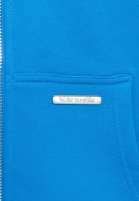 Blauer Sweatshirt-Stoff mit Fronttasche. Eigenschaften: silbernes Logo-Schild mit eingravierter Aufschrift "bazix republic" und Metallreißverschluss. Weiche Textur.