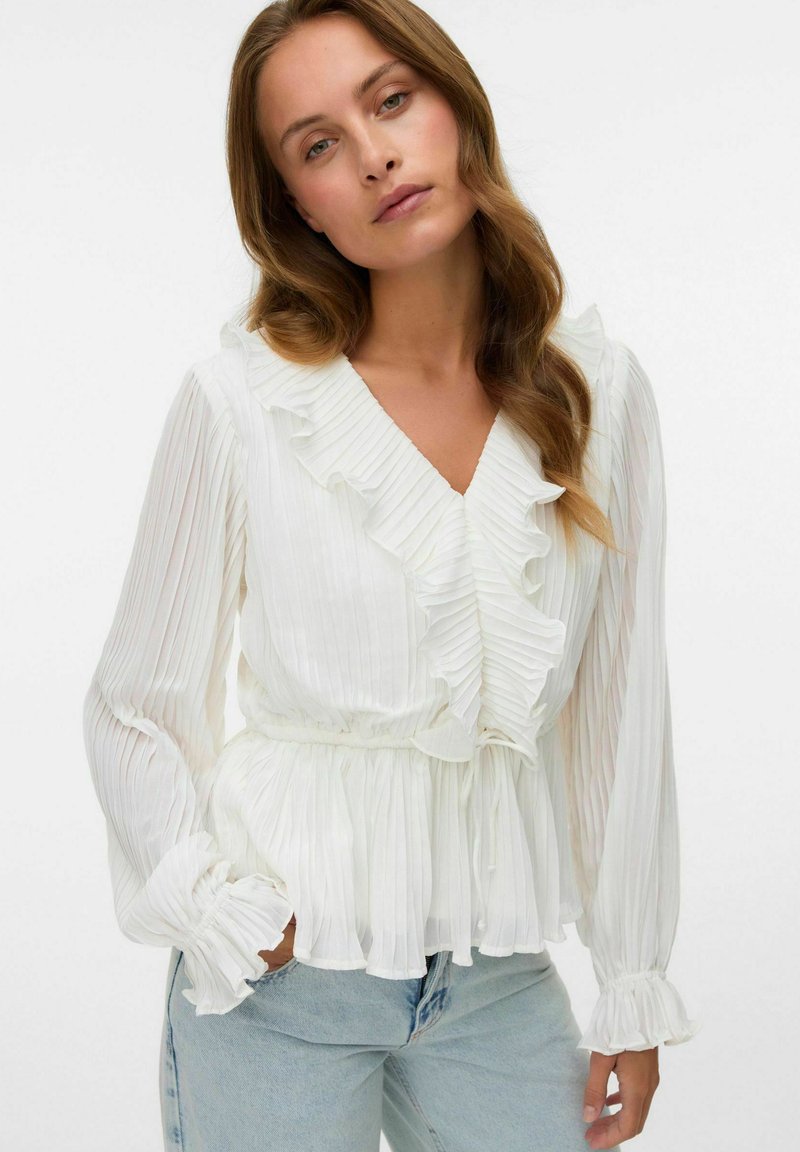 Hvit plissert bluse med volangkant ved hals og ermer, lange ermer, innsnevret midje og myk, teksturert stoff. Kombinert med lyseblå jeans.