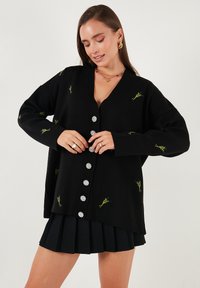 Crna pletena cardigan jakna s V-izrezom, ukrašena zelenim cvjetnim vezom i velikim srebrnim gumbima. Nosi se preko plisirane crne suknje.