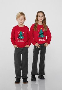 Rote gestrickte Pullover mit einem Grafik eines Weihnachtsbaums und dem Text "Rockin' Christmas". Ein Kind trägt schwarze Jeans und Schuhe und steht nebeneinander.