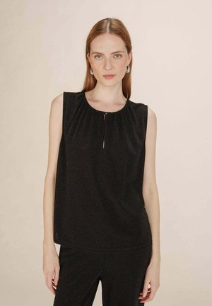 Femme à la peau claire et aux cheveux roux portant un top sans manches noir et un pantalon noir assorti, posant sur un fond beige.