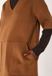 Gilet en mélange de laine marron avec col en V, doté de deux poches avant, manches longues foncées et une texture douce. Le design est minimaliste et moderne.