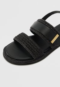 Sorte sandaler med to brede stropper - en glat læderstrop og en flettet strop. Tykt sål med en tekstureret kant og prægede logo detaljer på siden.