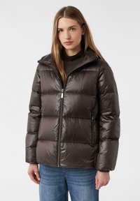 Bruine pufferjack met een glanzende afwerking, voorzien van een ritsluiting, hoge kraag en een gewatteerd ontwerp. Gedragen over een donkere top en blauwe spijkerbroek.