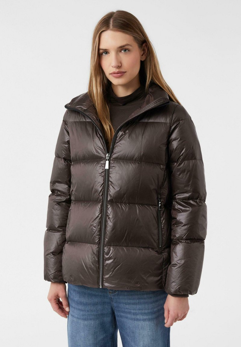 Bruine pufferjack met een glanzende afwerking, voorzien van een ritsluiting, hoge kraag en een gewatteerd ontwerp. Gedragen over een donkere top en blauwe spijkerbroek.