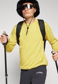 Cămașă galben deschis cu mâneci lungi, cu un model texturat, rucsac negru și pantaloni închiși la culoare cu logo-ul "TERREX". Accesoriile includ bețe de trekking și o pălărie neagră.