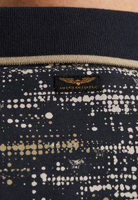 Marineblauw stoffen shirt met een geribde kraag, voorzien van een gestructureerd patroon van gouden en witte spikkels. Inclusief een zwart label met gouden tekst.
