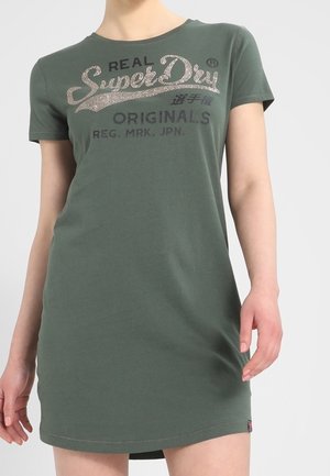 Kobieta nosząca oliwkową sukienkę z krótkim rękawem z napisem „Real SuperDry Originals” na piersi.