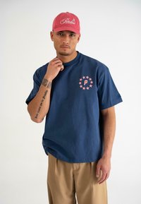 Mørkeblå bomulds T-shirt med korte ærmer, der har et pink rundt logo med stjerner. Båret sammen med beige bukser og en pink baseballkasket.